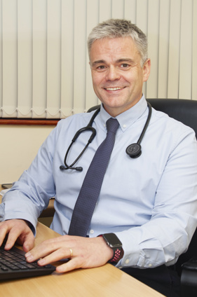 DR sitting at desk looking to camera.