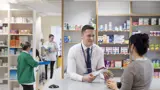 A smiling male pharmacist talking to a patient in a pharmacy with medicines stacked on the shelves in the background